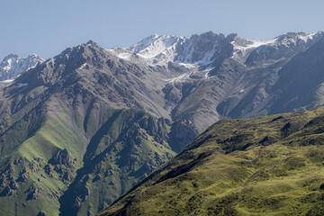 Fototapeta premium A rugged, greenery-covered mountain dominates the foreground while majestic snow-covered peaks rise behind it, all beneath a crisp azure sky