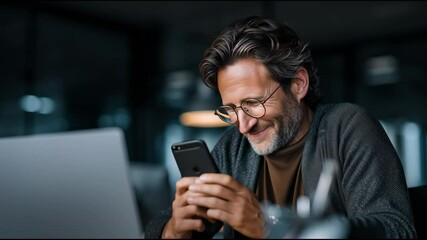 Happy businessman wearing glasses and a cardigan is using his smartphone while working late at night at his desk in a modern office with his laptop open - Powered by Adobe