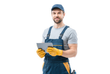 A smiling male worker in a gray t-shirt and blue overalls holds a tablet while wearing yellow gloves. standing against a white background. ready for a task in a professional setting