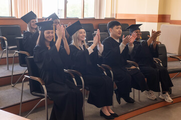 Young people in graduation gowns listen to a speech and applaud. 