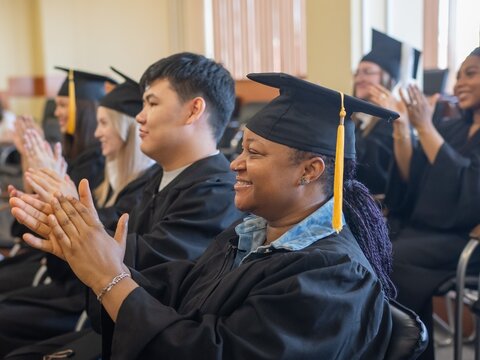 Young people in graduation gowns listen to a speech and applaud. 
