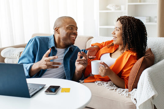Cheerful Black Couple Relaxing Together With Laptop And Coffee In Living Room, Happy Young African American Spouses Resting At Home, Having Hot Drinks, Using Computer And Chatting, Closeup Shot