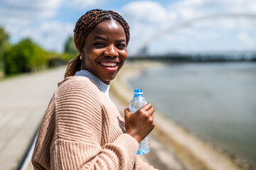 Portrait of cheerful young black woman enjoying sunny day in the city. She is holding bottle of...