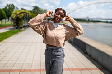 Happy young black woman is walking on foot path, listening music and smiling. She is enjoying sunny...