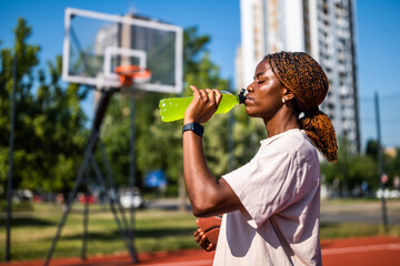 Young black woman practicing basketball alone at sports court in the morning. She is drinking...