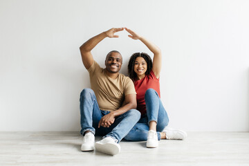 Happy young Black couple sitting on the floor forming roof shape with their arms, symbolizing home...