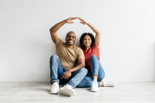 Happy young Black couple sitting on the floor forming roof shape with their arms, symbolizing home and future. Conceptual image of unity, protection, and love, copy space
