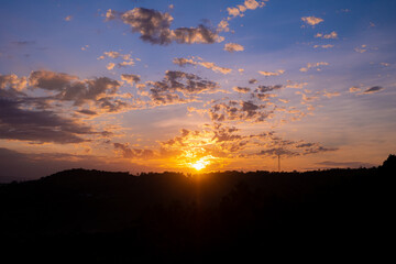 Vibrant Sunrise Over Hilly Terrain With Colorful Sky and Clouds. Golden sunlight illuminates a scenic hilly landscape while vivid orange and blue tones color the sky during sunrise.