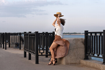 Wind lifts skirt as woman holds straw hat by river