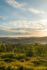 Beautiful sunlight illuminates a vast landscape of trees, grass, and misty mountains, creating a...
