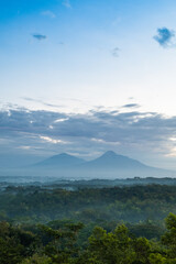 Fototapeta premium A breathtaking view of two majestic mountain peaks shrouded in mist, surrounded by lush green forest and under a dramatic cloudy sky, creating a serene and enchanting natural landscape.