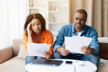 Upset black couple checking bills, sitting on couch, reading loan documents, unhappy african american man and woman holding papers, counting monthly spendings, planning family budget at home
