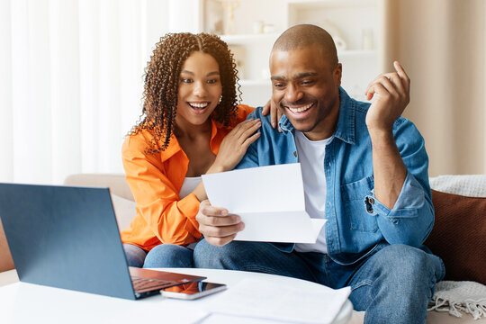 Surprised and smiling African American couple reading financial letter together at home, happy black female reacting positively while seated near laptop and documents, closeup
