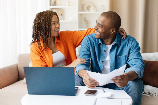 Happy African American couple smiling while reading papers at home, expressing joy and excitement over good news, black spouses surrounded by laptop and notebooks, closeup