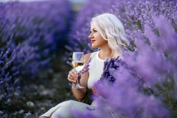 Blonde lavender field holds a glass of white wine in her hands. Happy woman in white dress enjoys lavender field picnic holding a large bouquet of lavender in her hands . Illustrating woman's picnic