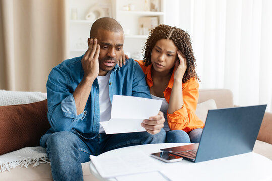 Young African American couple looking distressed as they read financial letter at home, black millennial spouses sitting close on couch, reacting to negative financial news