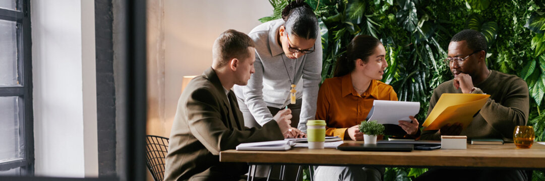 Website header shot of of diverse group of young adult and middle aged colleagues collaborating at table, reviewing documents and discussing business strategy in modern office with green plant wall