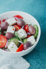 Greek salad with feta cheese olives cucumbers tomatoes and onions in white bowl on blue background