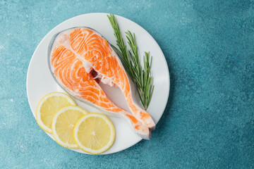 Raw salmon steak with lemon slices and rosemary on white plate over blue background