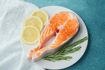Closeup raw salmon steak with lemon slices and rosemary on white plate over blue background