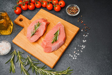 Raw veal steaks with rosemary on cutting board surrounded by cherry tomatoes oil salt and pepper