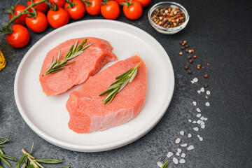 Raw veal steaks on white ceramic plate with tomatoes herbs salt oil and pepper