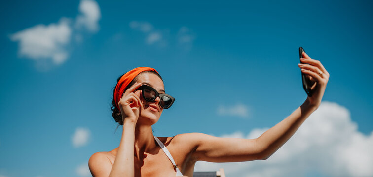 Young woman sitting by swimming pool outdoors in backyard garden, taking selfie.