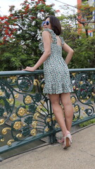 A confident Asian woman in a floral dress poses gracefully on a decorative bridge, surrounded by...