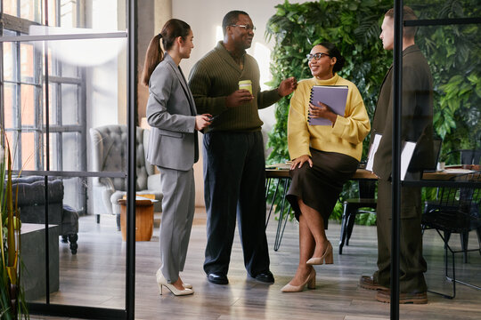 Group of young adult and middle aged multiethnic business professionals discussing project in modern office, Caucasian woman holding notebook, Black man holding coffee cup, teamwork concept