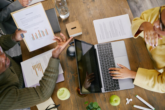 Overhead view of Black man presenting business charts to young adult Black woman working on laptop, both discussing financial data at wooden table with documents and office supplies