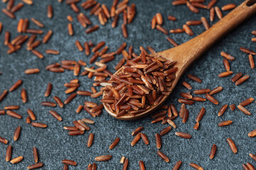 Wooden spoon filled with red rice on black background with scattered grains