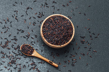 Black rice in wooden bowl with spoon and scattered grains on dark background