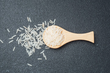 Wooden spoon filled with white rice and scattered grains on dark surface