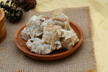 A plate of steamed dumplings served with a side of dipping sauce, placed on a burlap cloth with pinecones in the background.