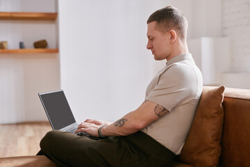 Side view of focused Caucasian young adult man sitting on sofa typing on laptop while working at...