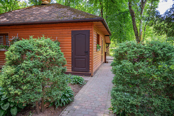 A rustic wooden cabin with a brown door stands amidst lush green bushes and trees, with a paved walkway leading up to it.