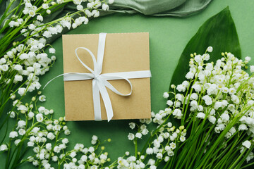 Gift box with white ribbon surrounded by lily of the valley on green background