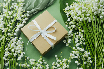 Gift box with white ribbon surrounded by lily of the valley on green background
