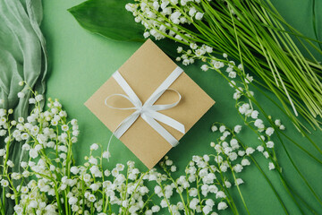 Gift box with white ribbon and lily of the valley flowers on green background