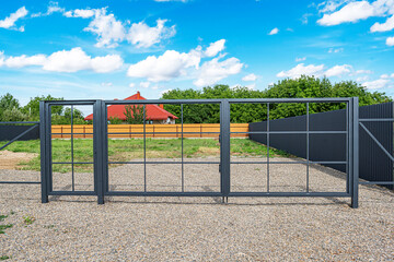 A modern metal gate with a grid design stands closed on a gravel driveway, leading to a fenced...