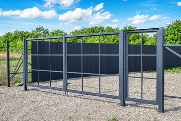 A modern metal gate with a grid design stands closed on a gravel driveway, leading to a fenced...