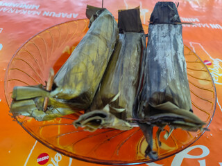Three packets of lontong nasi wrapped in yellowish green banana leaves and tied with a stick,...