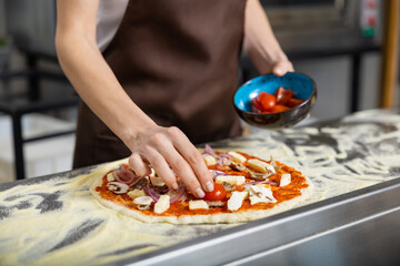 Cooker in apron working in the pizzeria kitchen and putting topping on the pizza