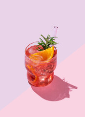 Red orange cocktail or lemonade with ice and rosemary on a pink background with shadows.