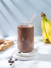 Chocolate coffee smoothie with banana in a glass on a white blue background with morning shadow.