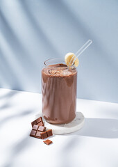 Chocolate smoothie with banana in a glass on a white blue background with morning shadow.