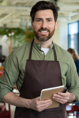 Young bearded man in apron looking confident while waiting for the client in pizzeria