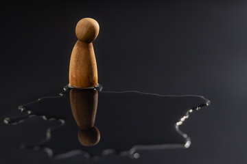 A wooden peg doll stands alone, staring down at its reflection in a puddle. Symbolizes symbolizing mental health crisis, deep depression, stress, and emotional pressure