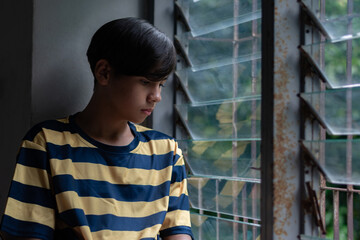 A teenage boy in a yellow and navy striped shirt sits quietly by an old window, looking down with a thoughtful expression. Captures a mood of solitude and reflection.