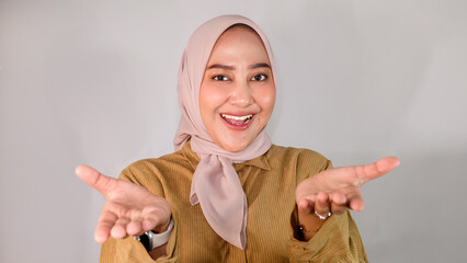 Beautiful young Asian Muslim woman wearing hijab and brown shirt conveying idea while smiling and stretching both hands forward on isolated white background.